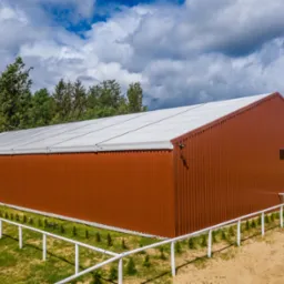 Gotowa hala z brązowej blachy trapezowej z białym dachem, otoczona zielenią i białym ogrodzeniem, widok z góry, błękitne niebo z chmurami.