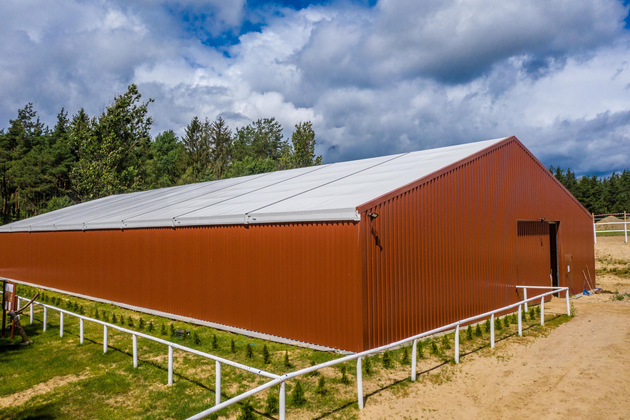 Gotowa hala z brązowej blachy trapezowej z białym dachem, otoczona zielenią i białym ogrodzeniem, widok z góry, błękitne niebo z chmurami.