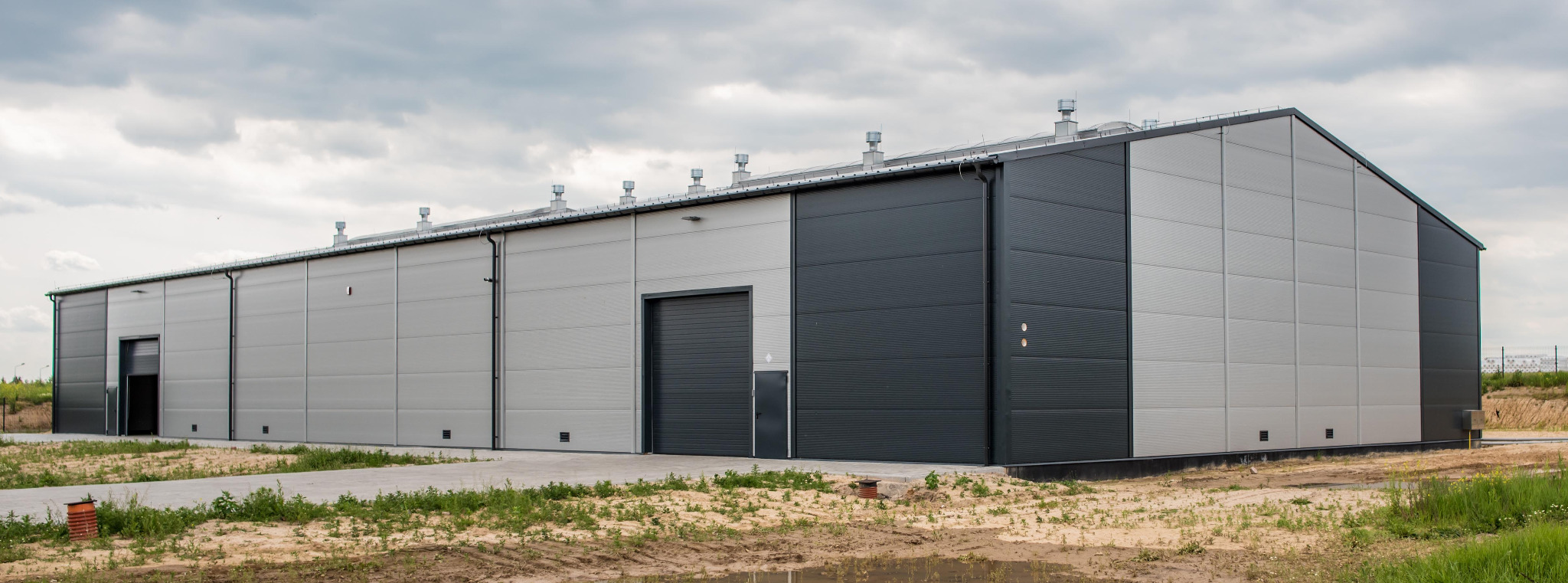 Nowoczesna hala magazynowa z szarej blachy trapezowej, z dwoma bramami segmentowymi i systemem wentylacji na dachu, otoczona terenem zielonym po deszczu.