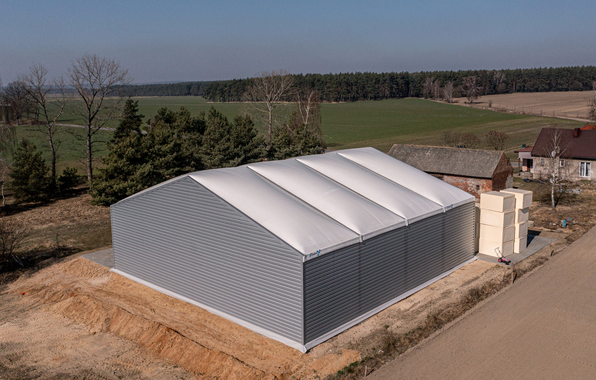 Nowoczesna hala magazynowa z szarej blachy falistej i białym dachem membranowym, widok z lotu ptaka, w tle pola i las.