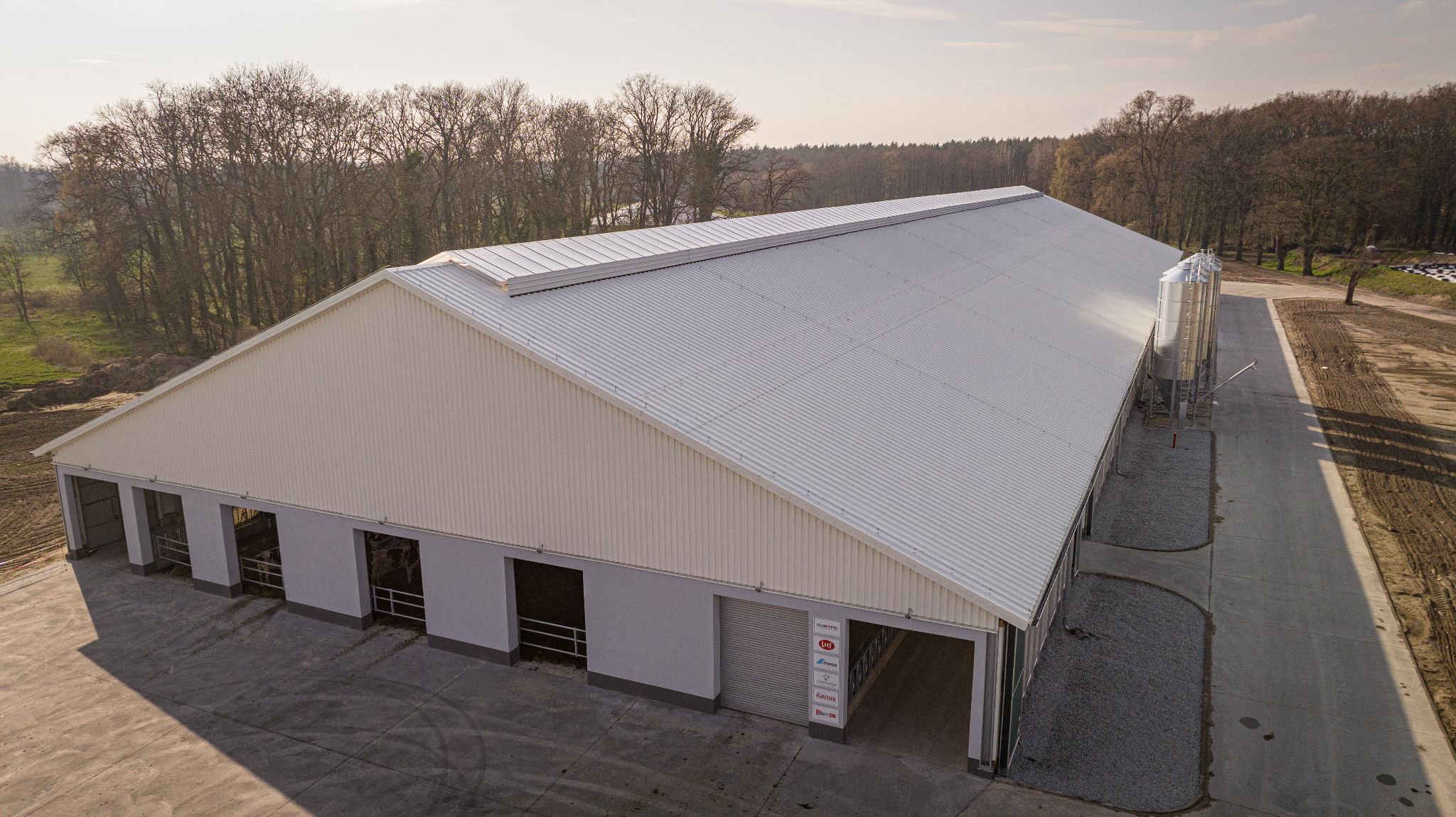 Widok z góry na dużą halę z białymi ścianami i srebrnym dachem z blachy trapezowej, przylegającą do niej betonową drogą i silosami, otoczoną drzewami bez liści.