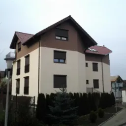 Dwukondygnacyjny dom z brązowo-białą elewacją, czerwoną dachówką i zewnętrznymi roletami, widziany z poziomu ulicy w pochmurny dzień, z widocznym fragmentem chodnika i ogrodzenia.