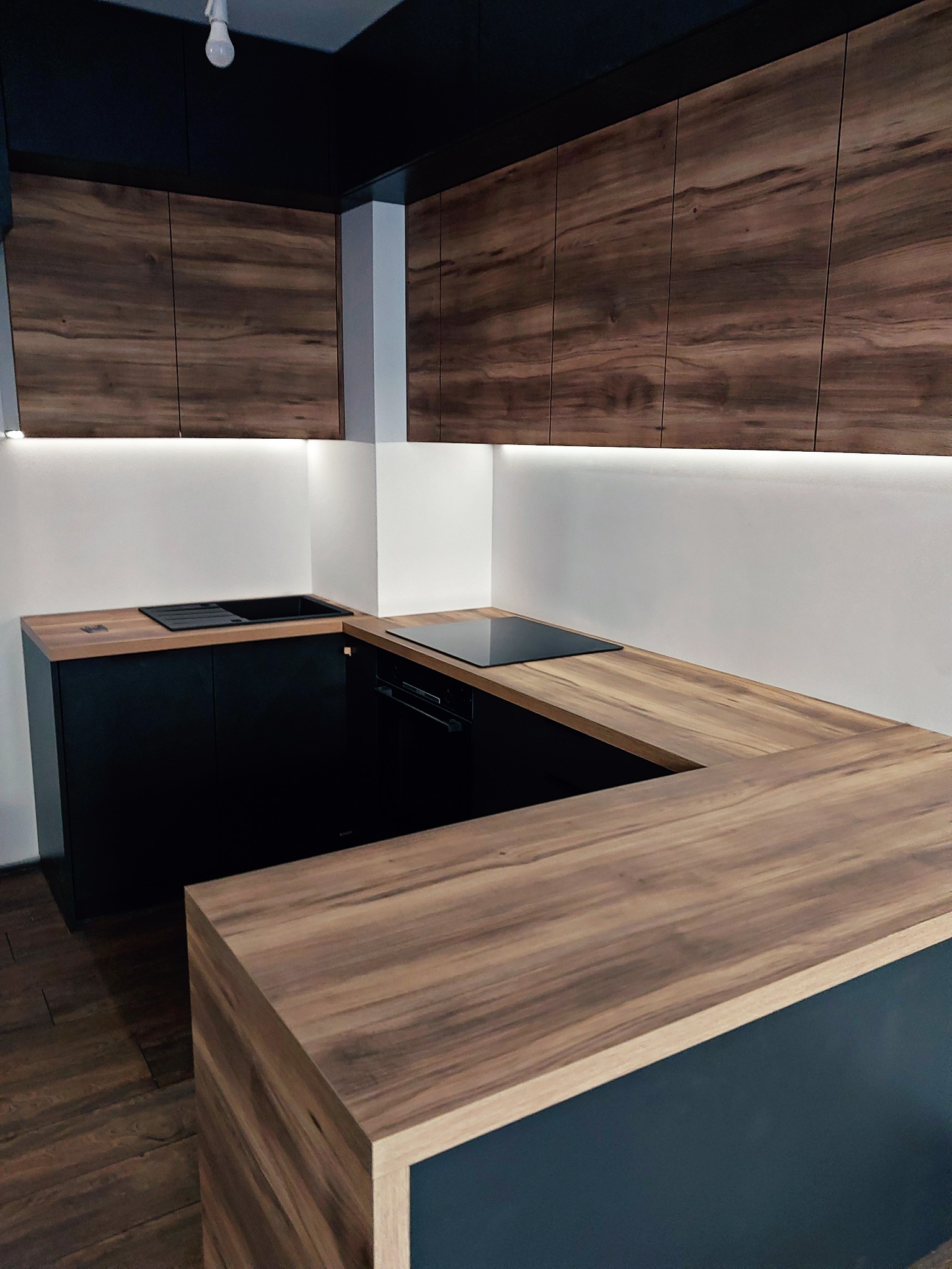 Nowoczesna kuchnia z drewnianymi szafkami górnymi i blatem w kształcie litery U, czarne szafki dolne, oświetlenie LED pod szafkami górnymi, czarna płyta indukcyjna i zlew.