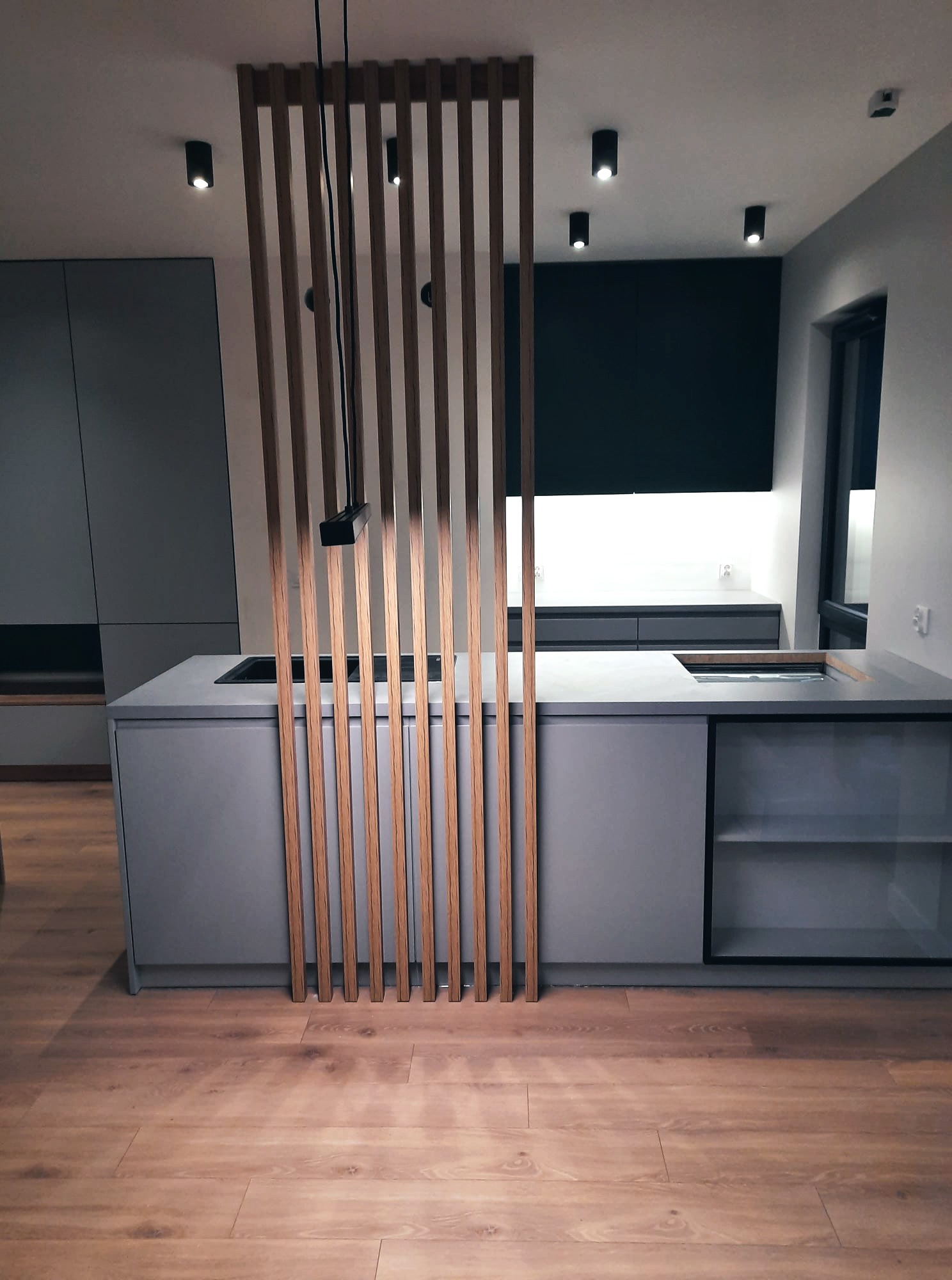 Nowoczesna kuchnia z szarymi szafkami, drewnianymi lamelami oddzielającymi przestrzeń i drewnianą podłogą, oświetlona punktowymi lampami.