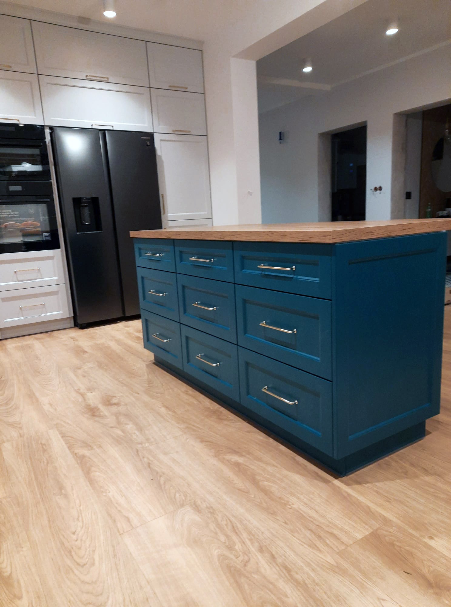 Wyspa kuchenna z drewnianym blatem i frontami w kolorze ciemnego turkusu, z metalowymi uchwytami, na tle białych szafek i czarnej lodówki side-by-side.