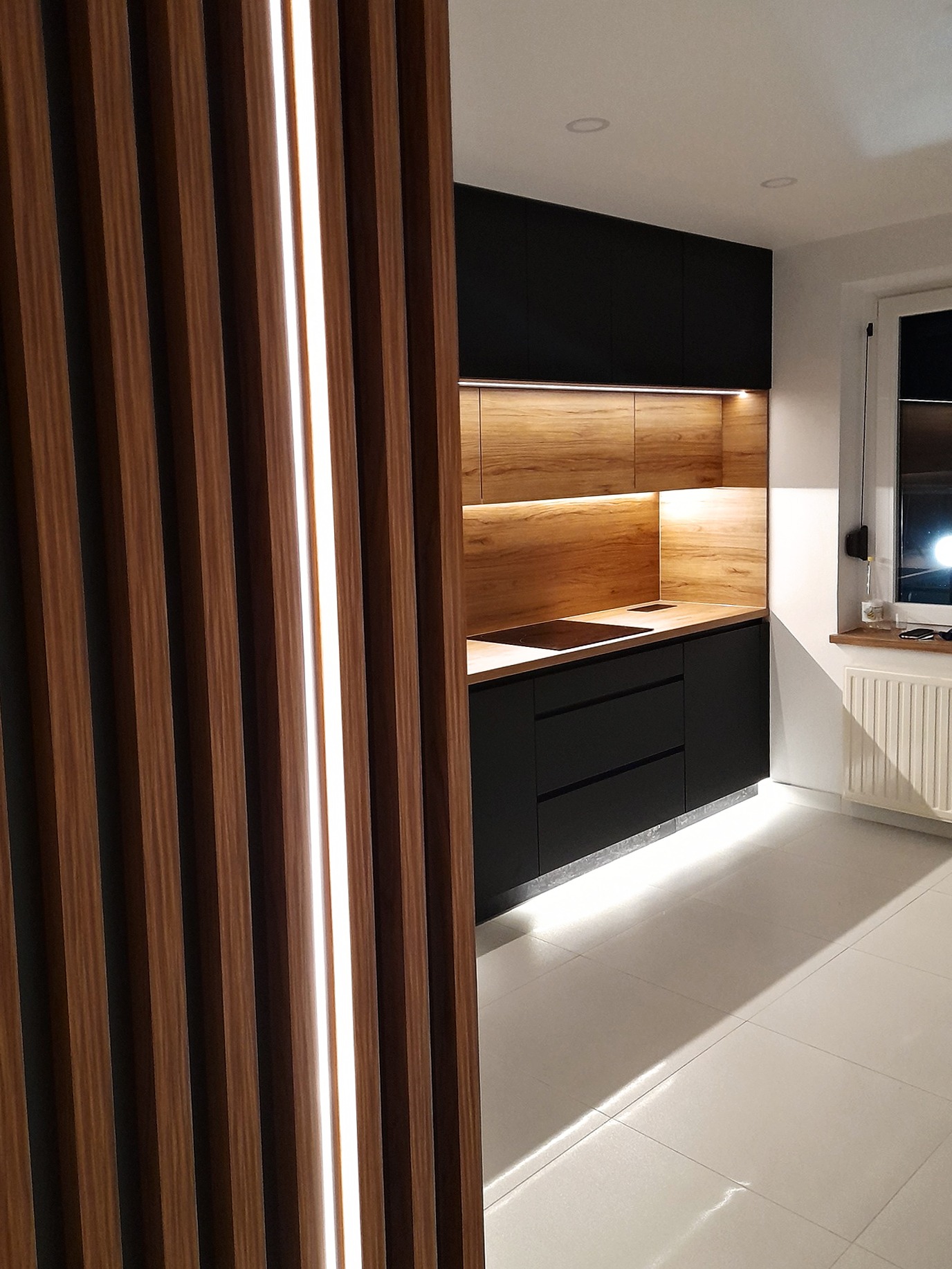 Nowoczesna kuchnia z czarnymi szafkami i drewnianymi akcentami, oświetlona listwami LED, widoczna przez dekoracyjną ściankę działową z pionowych lameli.