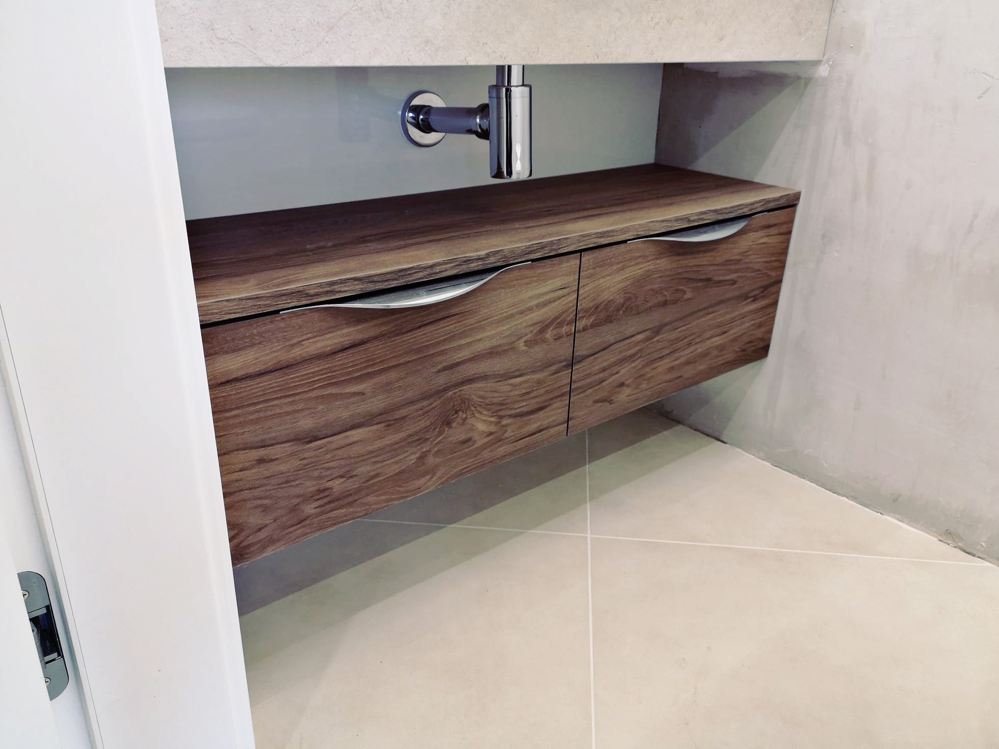 Szafka pod umywalkę z drewnopodobnym frontem i chromowanymi uchwytami, widoczny syfon i beżowe płytki podłogowe.