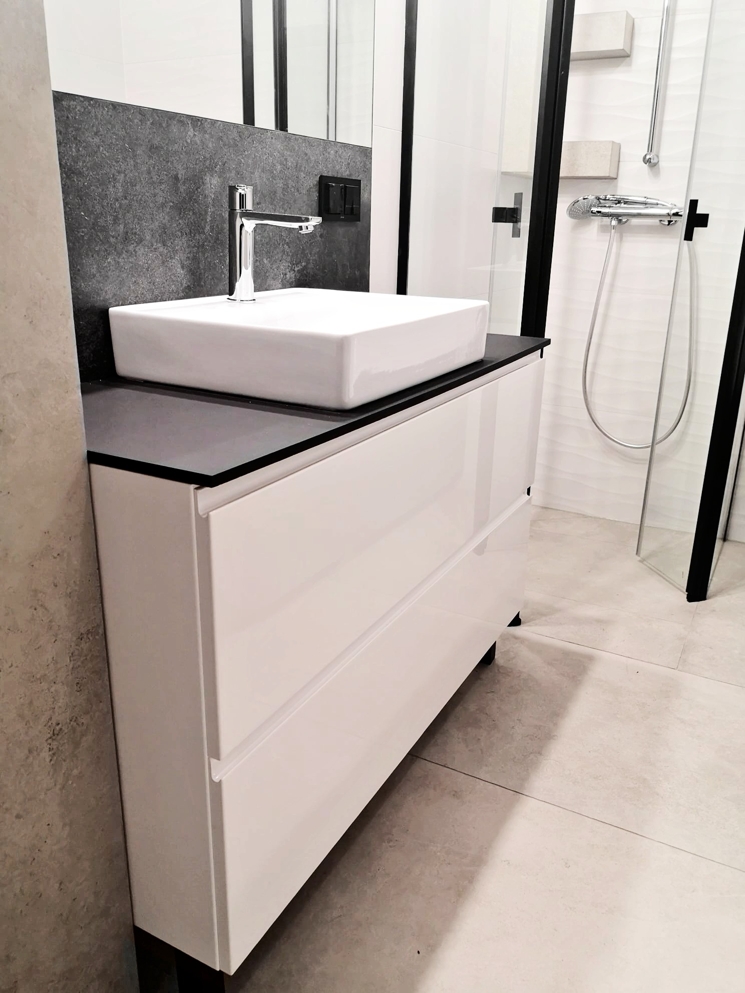 Nowoczesna łazienka z białą szafką pod umywalkę, czarnym blatem i kwadratową umywalką, widoczny fragment kabiny prysznicowej z czarnymi ramami.