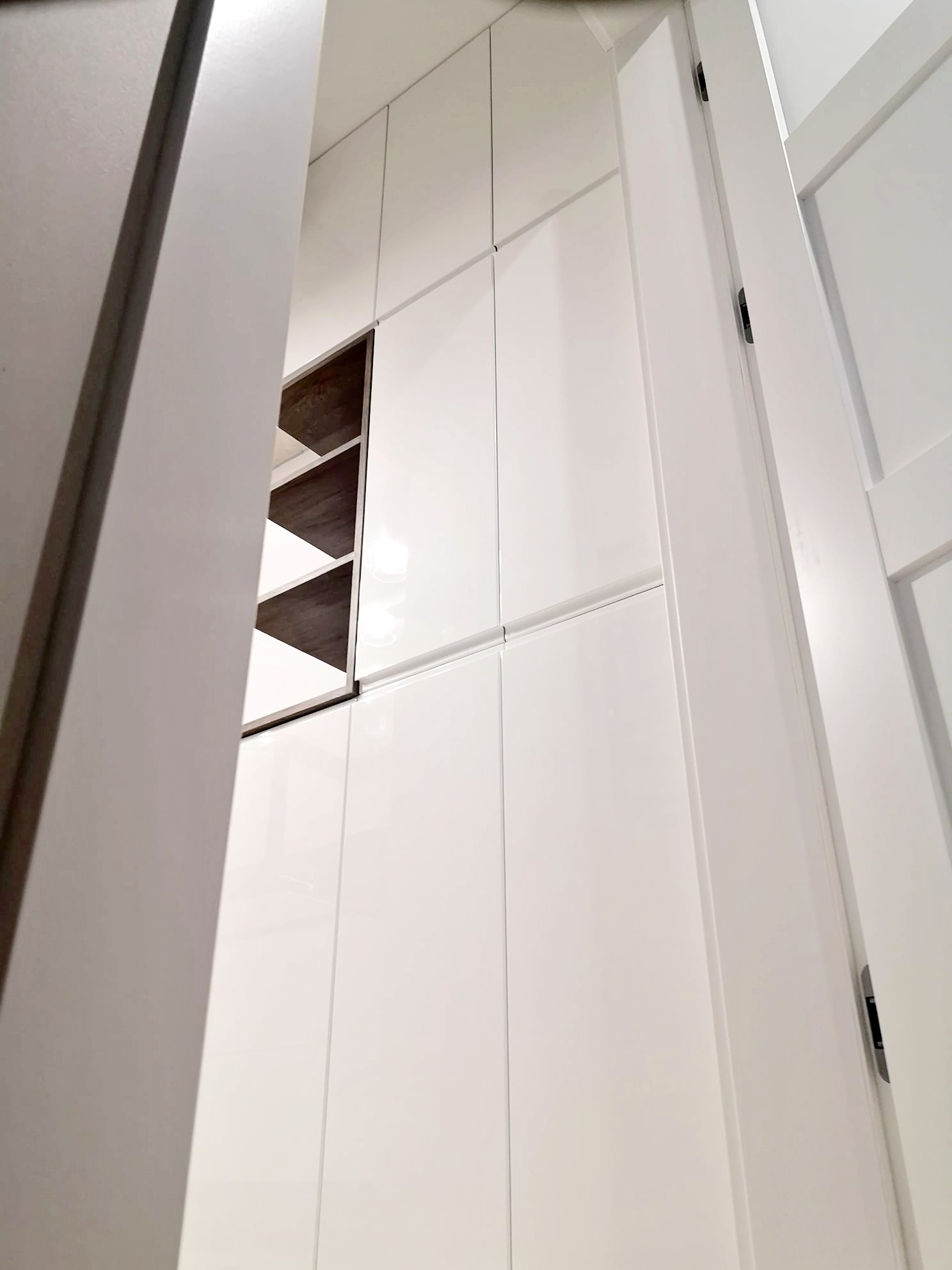 Wnętrze szafy wnękowej z białymi, lakierowanymi frontami i otwartą półką z ciemnego drewna, widziane z perspektywy wnętrza pokoju przez częściowo otwarte drzwi.