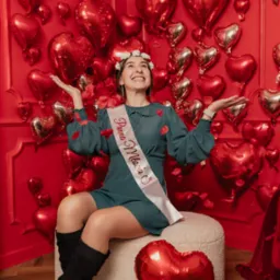 Uśmiechnięta kobieta w sukience i szarfie 'Panna Młoda' siedzi na pufie, otoczona czerwonymi balonami w kształcie serc i płatkami róż, patrząc w górę.