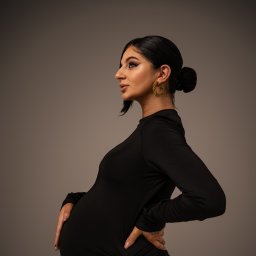 KARO Karolina Brzezińska - Kobieta w czarnej sukience, profil, ciążowy brzuszek, ręce na biodrach, kolczyki koła, upięte włosy, neutralne tło, elegancki makijaż.