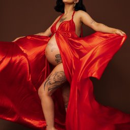 KARO Karolina Brzezińska - Kobieta w ciąży, z tatuażami, pozuje w czerwonej, satynowej sukni na brązowym tle. Rozpostarte ręce podkreślają krój sukni i uwydatniają ciążowy brzuszek.