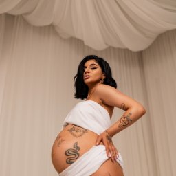 KARO Karolina Brzezińska - Kobieta w ciąży, owinięta białą tkaniną, prezentuje tatuaże na brzuchu i nogach na tle beżowej ściany i białego, drapowanego materiału.