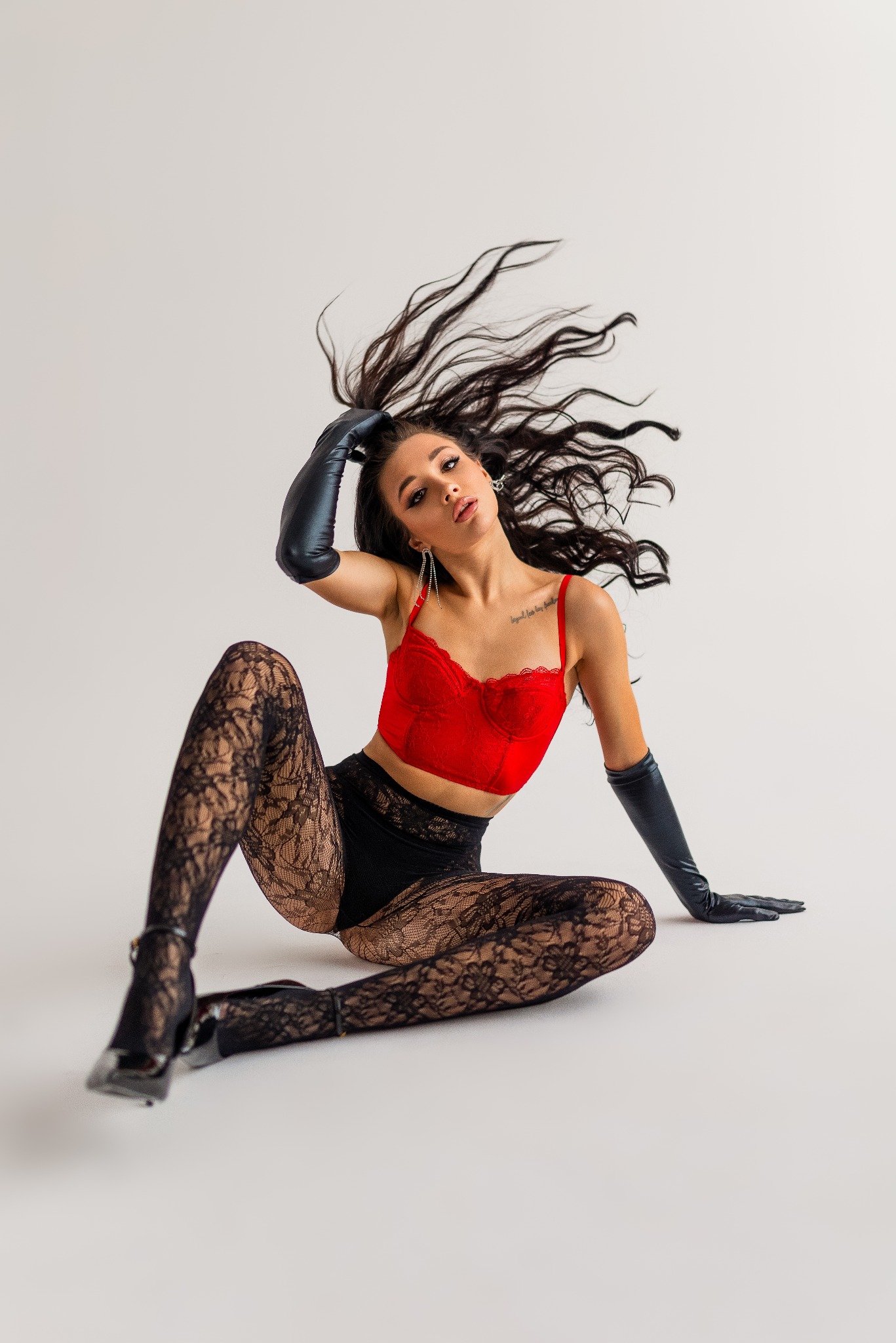 Kobieta w czerwonym koronkowym biustonoszu, czarnych rajstopach w kwiaty i długich skórzanych rękawiczkach, pozuje siedząc na podłodze z rozrzuconymi włosami i spojrzeniem skierowanym w górę.