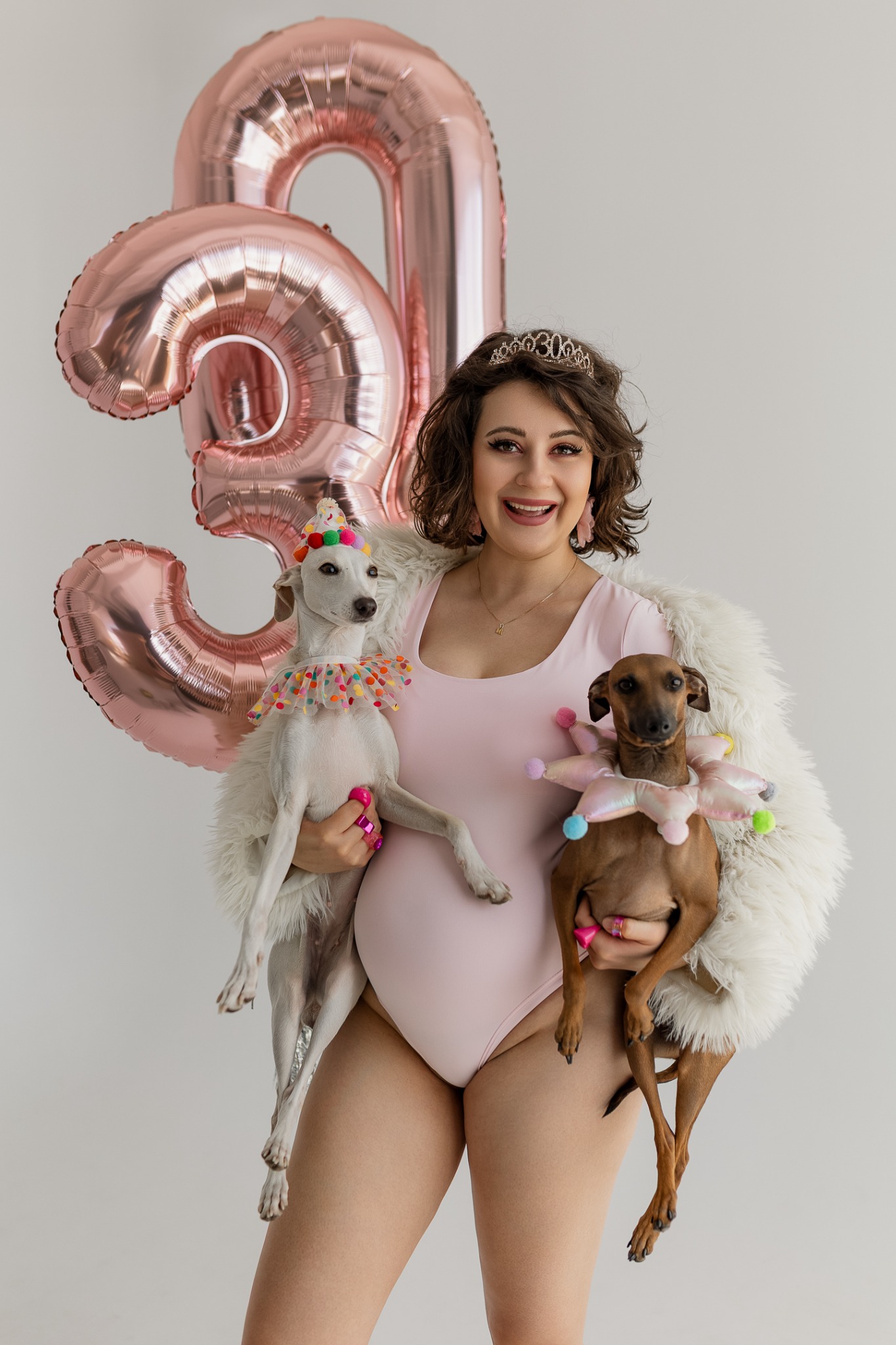 Uśmiechnięta kobieta w różowym body i futrze, trzymająca dwa psy w urodzinowych strojach na tle balonów w kształcie cyfry 30.