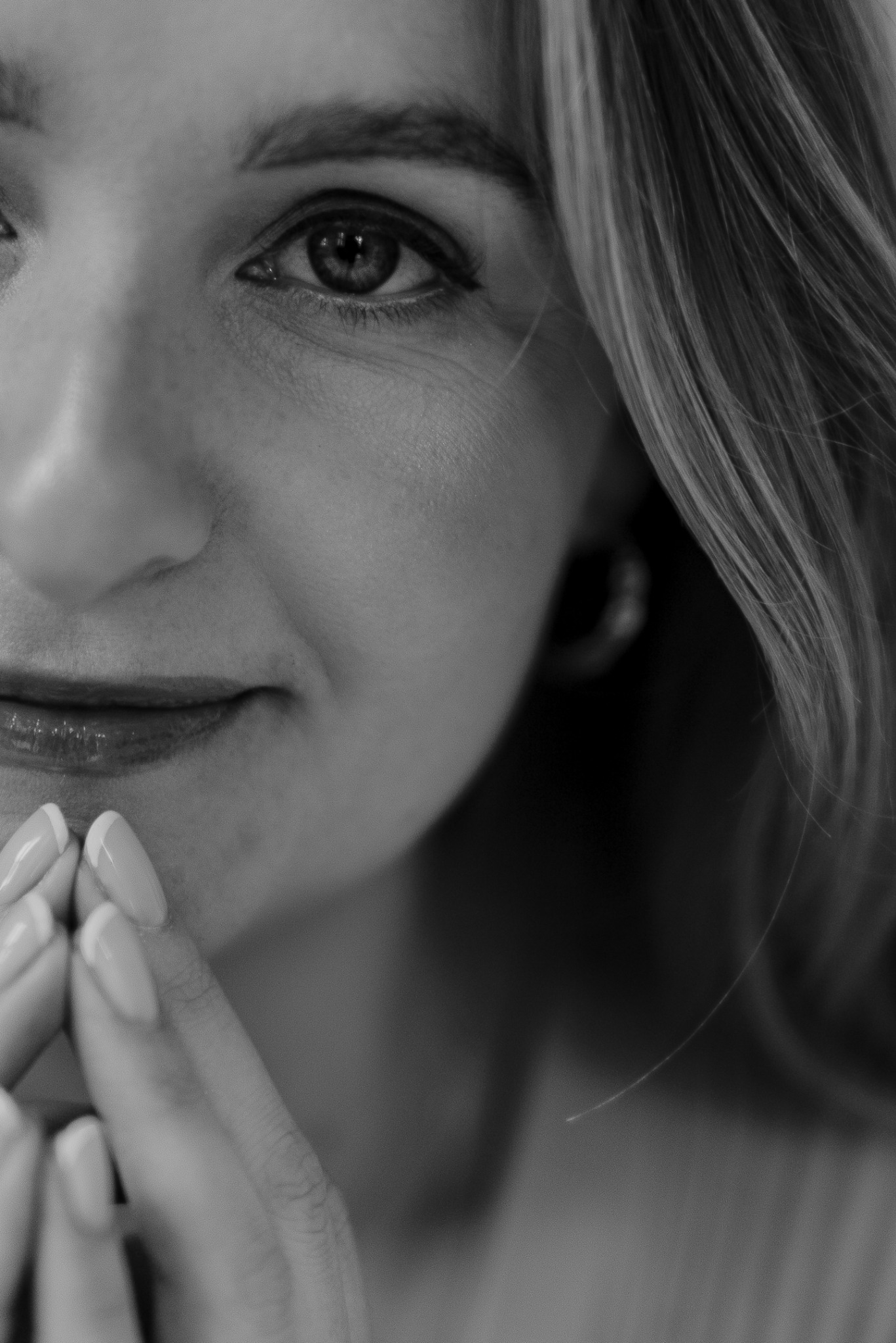 Portret zbliżeniowy kobiety w czerni i bieli. Widoczna część twarzy, w tym oko z delikatnym makijażem, usta i dłonie z jasnym manicurem. Subtelne oświetlenie.