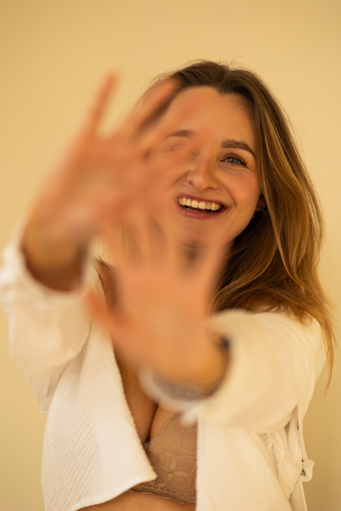 Uśmiechnięta kobieta w białym szlafroku zasłaniająca twarz dłońmi, z widocznym fragmentem biustonosza, na jasnym tle. Fotografia portretowa.