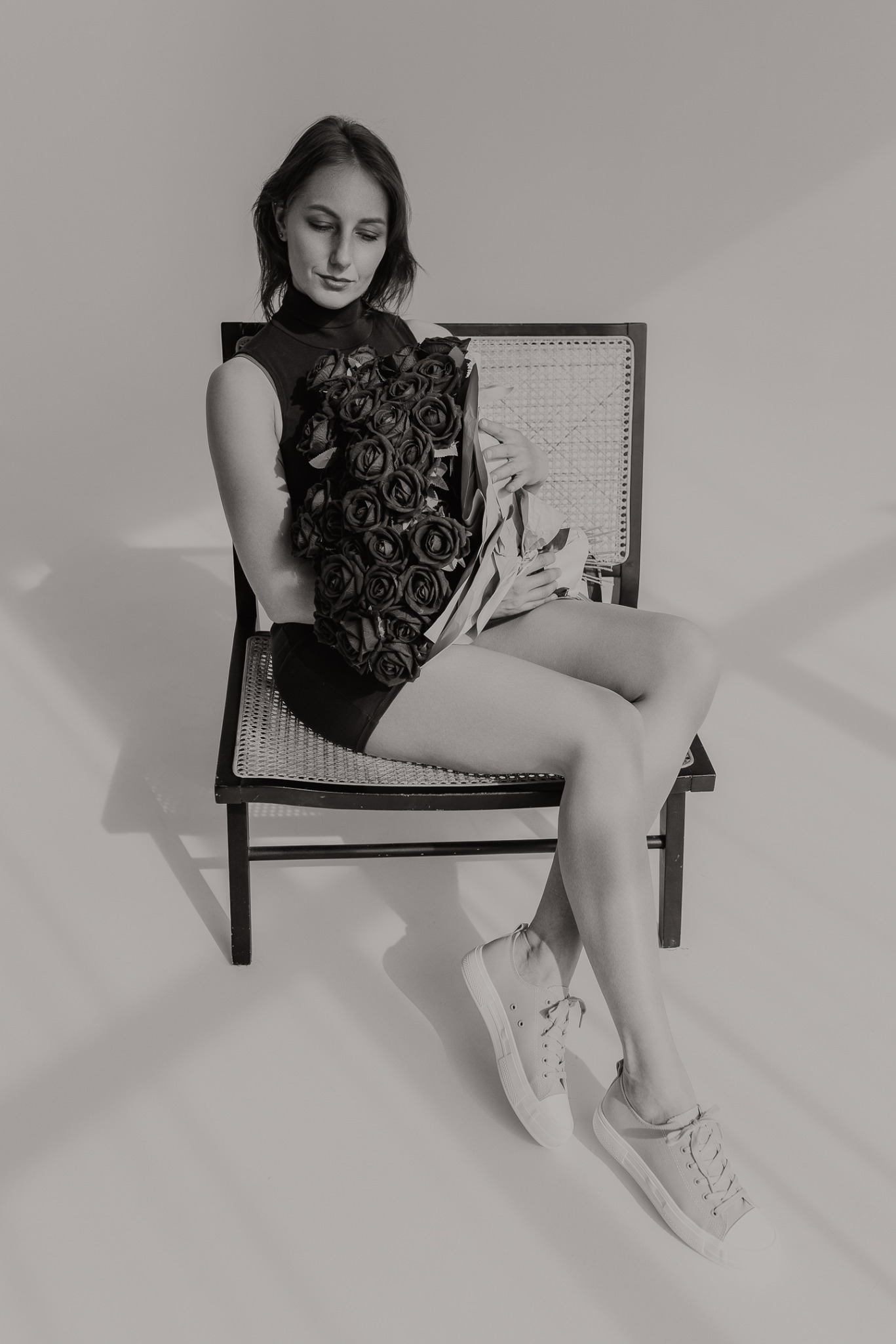 Kobieta siedzi na krześle, trzymając bukiet ciemnych róż. Ubrana w czarny top i szorty, ma na sobie jasne trampki. Minimalistyczne tło, monochromatyczna tonacja.