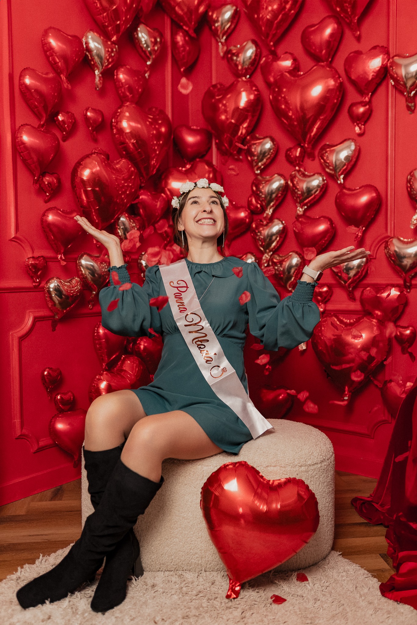 Uśmiechnięta kobieta w sukience i szarfie 'Panna Młoda' siedzi na pufie, otoczona czerwonymi balonami w kształcie serc i płatkami róż, patrząc w górę.