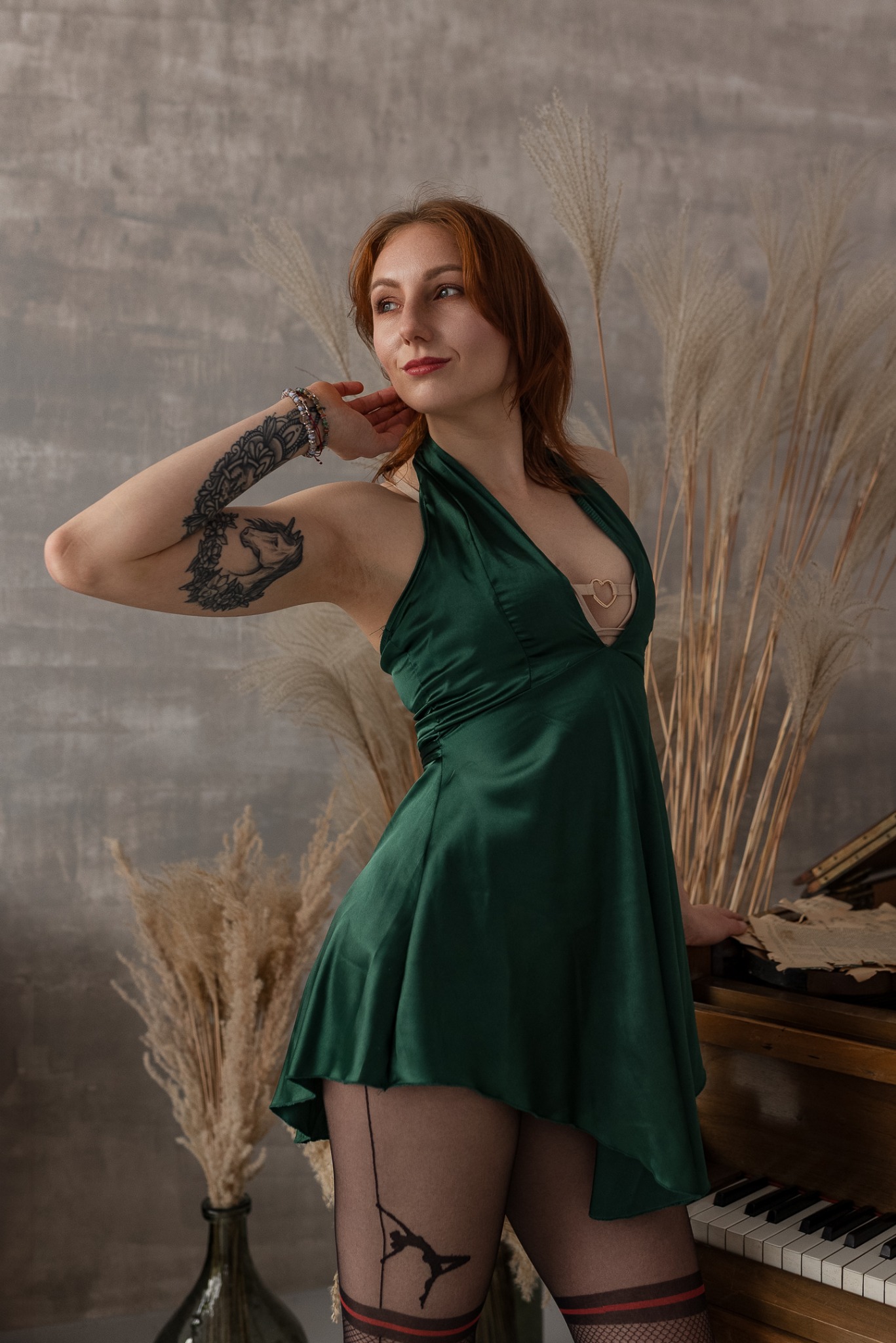 Kobieta w zielonej sukience pozuje przy pianinie w Grodzisku Mazowieckim. Widoczny tatuaż na ramieniu i rajstopy z nadrukiem. W tle dekoracja z traw.
