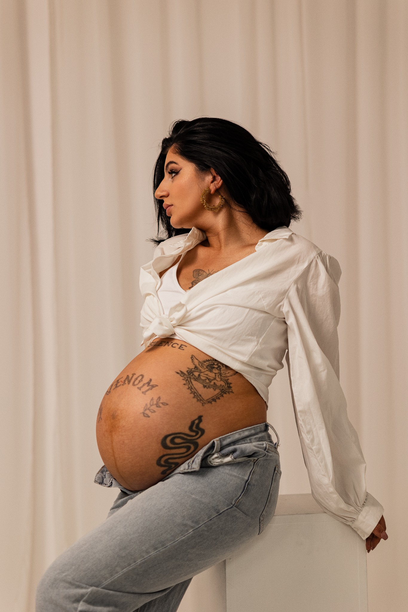 Kobieta w ciąży, z tatuażami na brzuchu, pozuje w białej koszuli i jeansach na tle jasnej zasłony. Widok z profilu, złote kolczyki koła, czarne włosy.