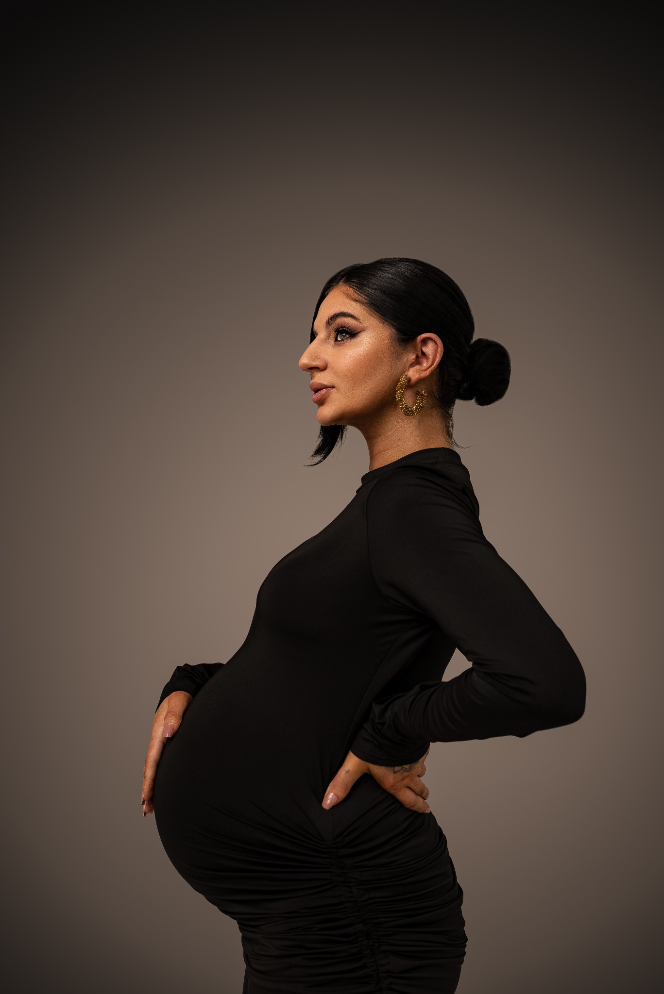 Kobieta w czarnej sukience, profil, ciążowy brzuszek, ręce na biodrach, kolczyki koła, upięte włosy, neutralne tło, elegancki makijaż.