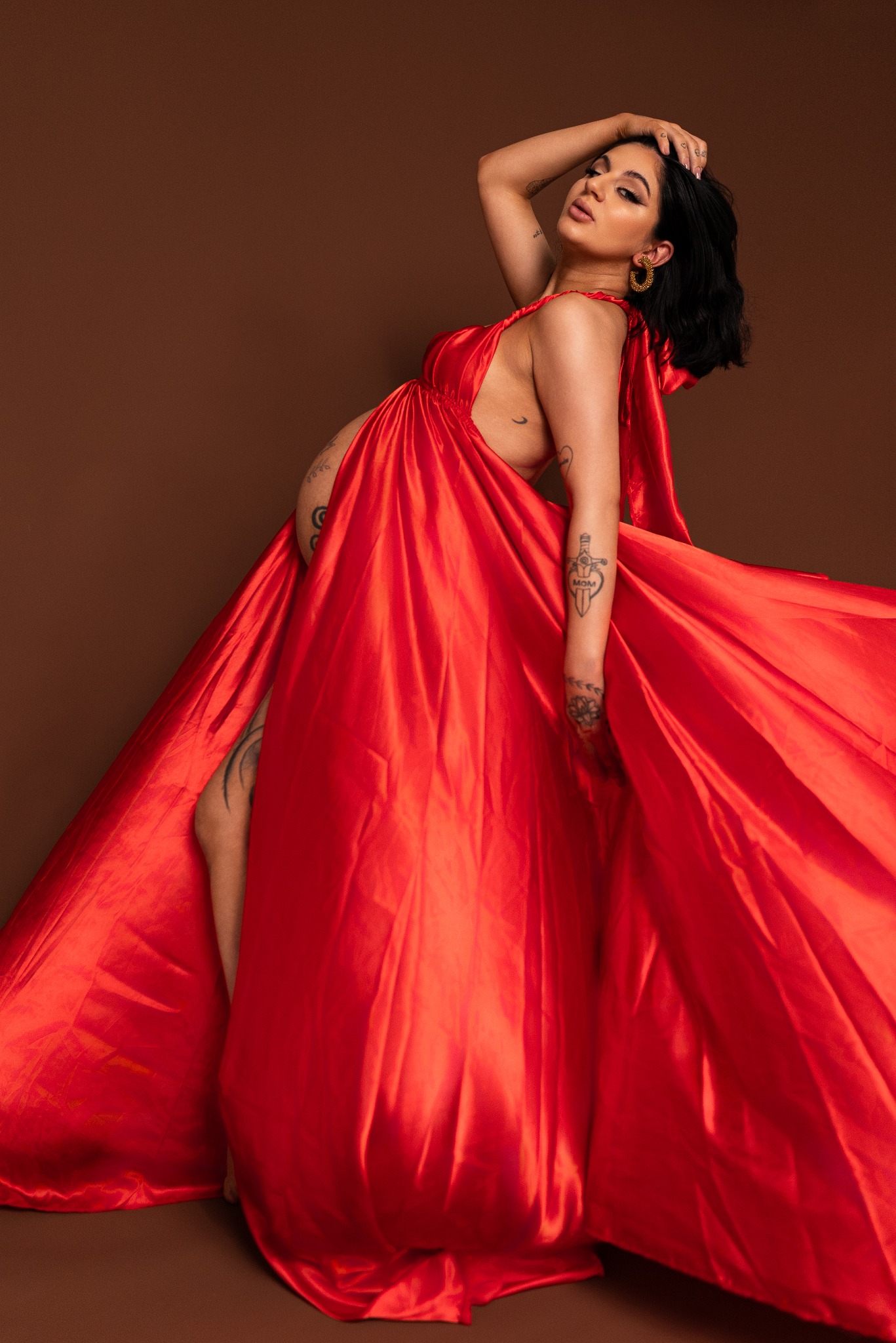 Kobieta w czerwonej, powłóczystej sukni, w dynamicznej pozie, z ręką na głowie. Widoczne tatuaże na ramionach i brzuchu. Ciemne włosy, złote kolczyki. Brązowe tło.