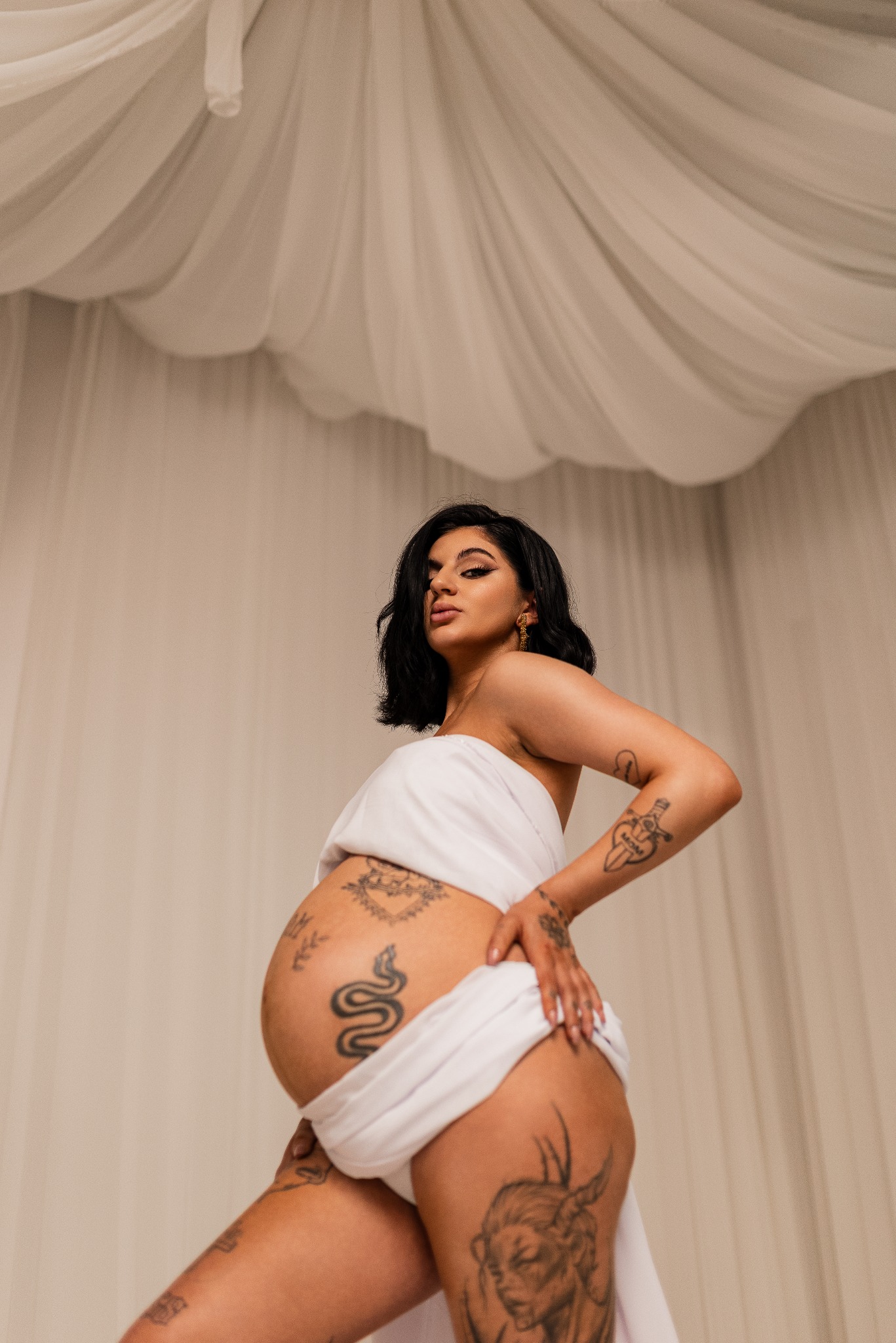 Kobieta w ciąży, owinięta białą tkaniną, prezentuje tatuaże na brzuchu i nogach na tle beżowej ściany i białego, drapowanego materiału.
