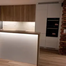 Nowoczesna kuchnia: połączenie drewnianych szafek górnych, białych szafek dolnych z wbudowanymi piekarnikami i oświetlonym blatem; ceglana kolumna w tle.