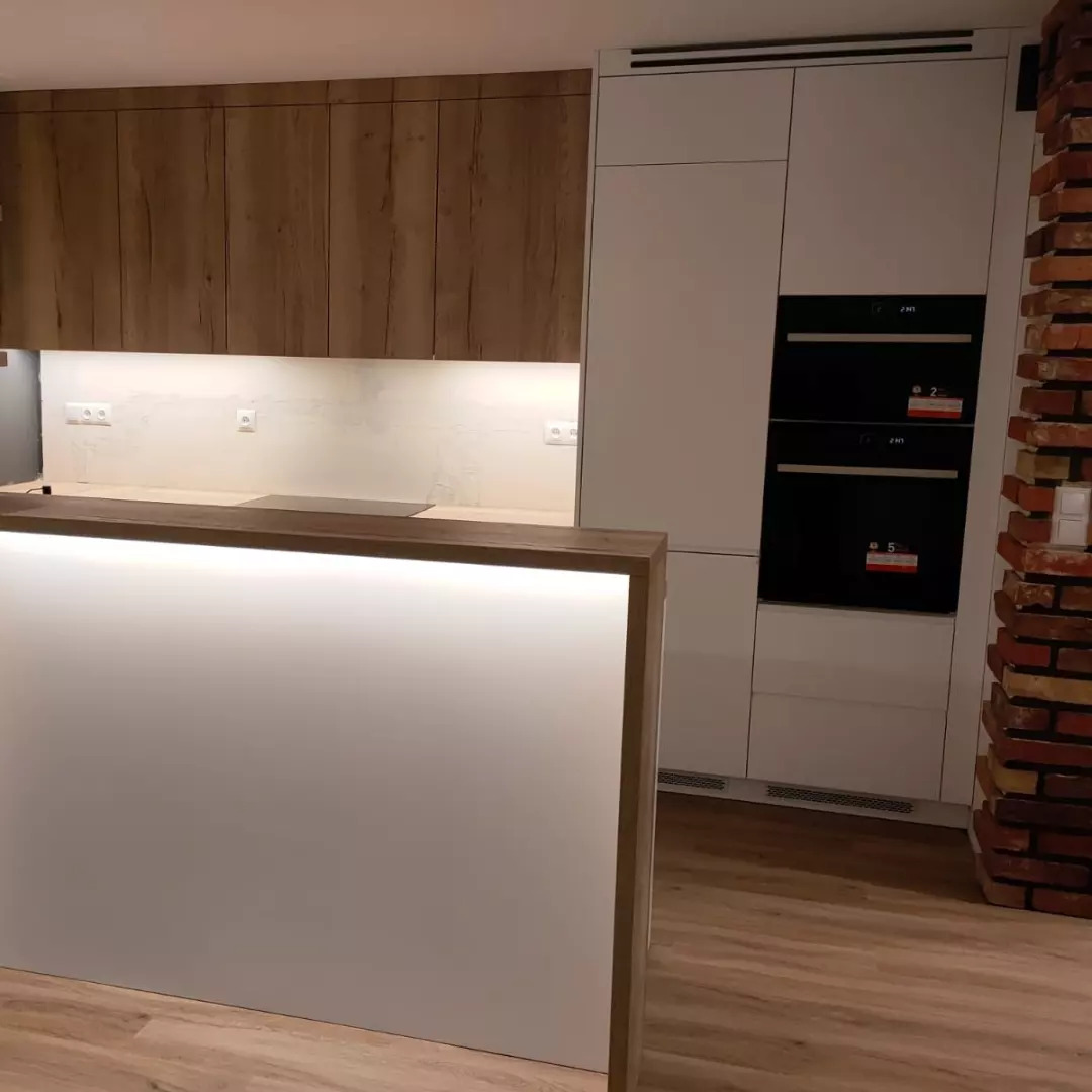 Nowoczesna kuchnia: połączenie drewnianych szafek górnych, białych szafek dolnych z wbudowanymi piekarnikami i oświetlonym blatem; ceglana kolumna w tle.