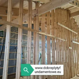 Wnętrze domu w trakcie budowy: drewniana konstrukcja szkieletowa ścian i stropu, widoczne izolacje i membrany, surowe drewno, częściowo oświetlone naturalnym światłem wpadającym przez okno.