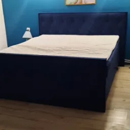 Niebieskie łóżko tapicerowane z pikowanym zagłówkiem, białą pościelą i drewnianą podłogą, widoczne oświetlenie sufitowe i obraz na ścianie.