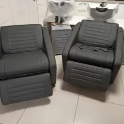 Dwa ciemnoszare, skórzane fotele fryzjerskie z regulowanymi podnóżkami, widoczne w salonie fryzjerskim, jeden z przewodami i kluczem na siedzisku.