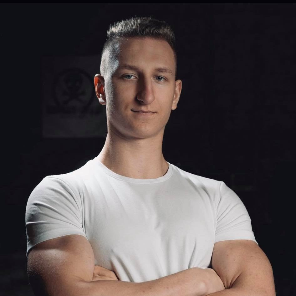 Młody mężczyzna w białym t-shircie, z założonymi rękami, na ciemnym tle z delikatnym logo czaszki w tle.