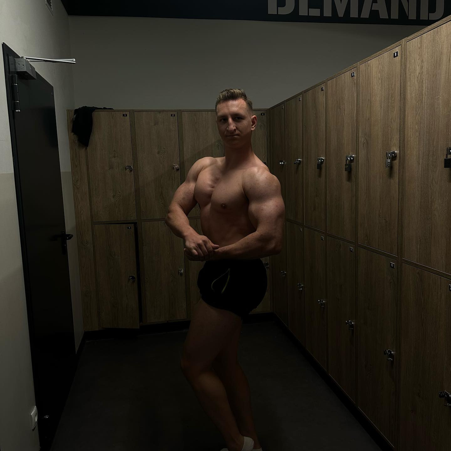 Mężczyzna prezentuje muskulaturę w szatni siłowni w Starej Iwicznej, pozując na tle rzędu szafek z numerami.