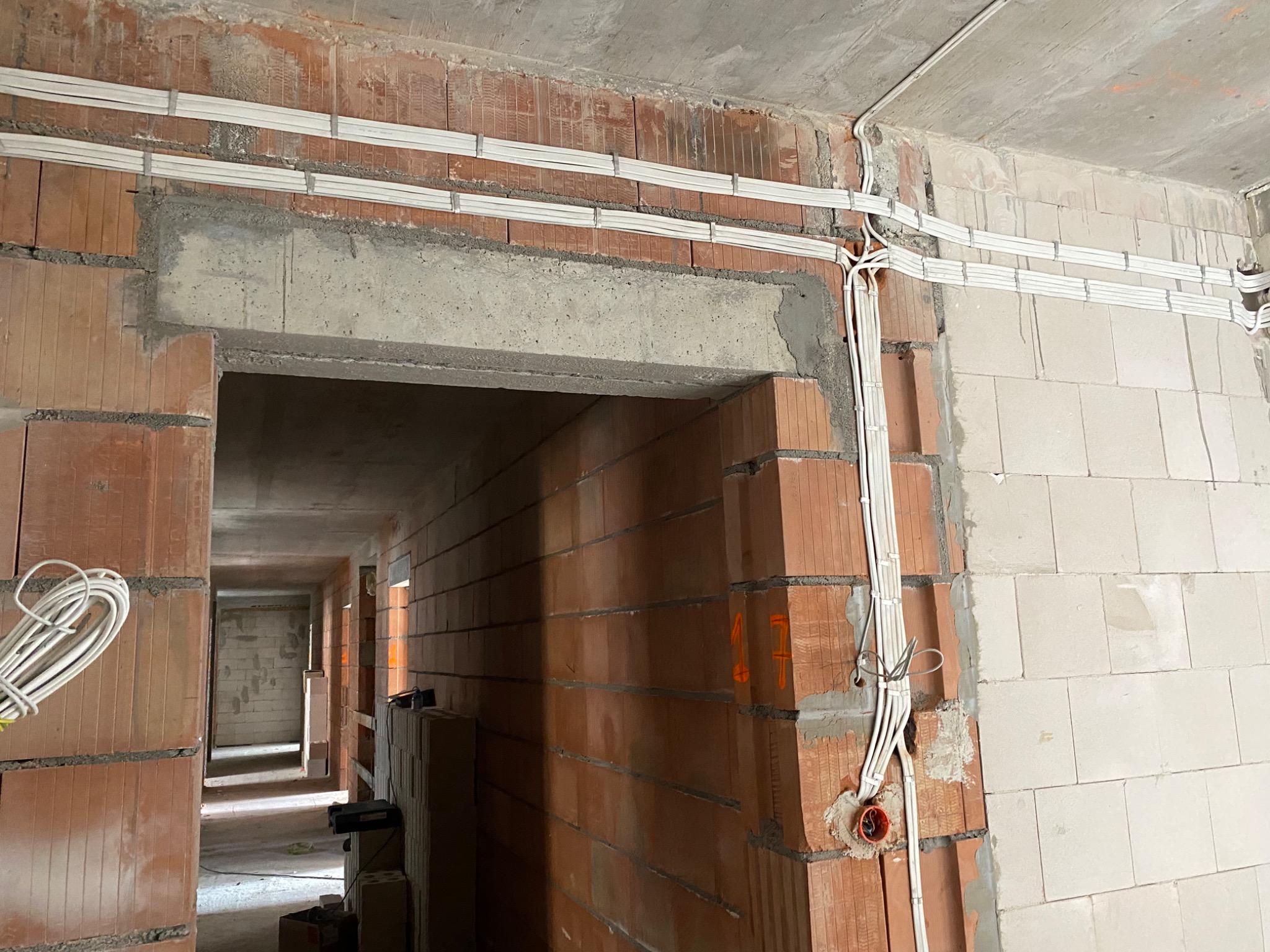 Surowy stan wnętrza w trakcie prac: widoczne przewody elektryczne prowadzone po ścianach z czerwonej cegły i betonu, zamocowane w listwach, z puszkami instalacyjnymi w ścianie.