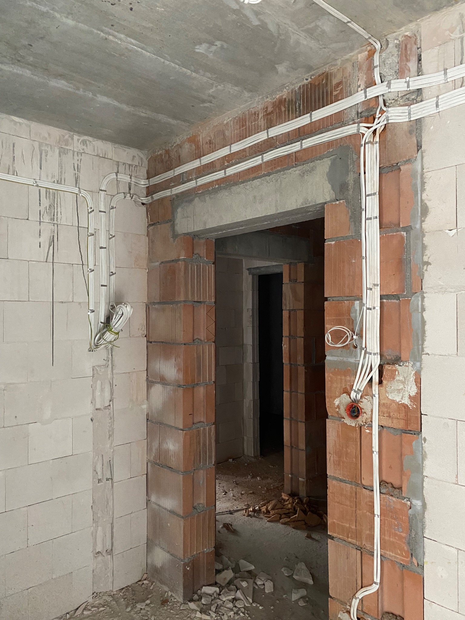 Surowy stan wnętrza w trakcie rozprowadzania instalacji elektrycznej: widoczne kable przymocowane do ścian z bloczków, surowy betonowy strop, otwór drzwiowy w stanie budowy.