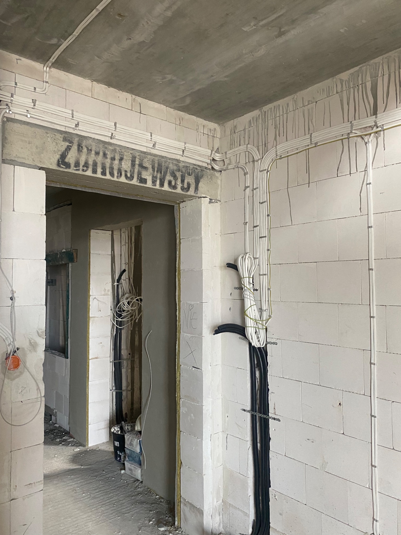 Surowy stan wnętrza w trakcie remontu: widoczne kable elektryczne prowadzone po ścianach z betonu komórkowego, częściowo zamaskowane w peszlach, z napisem 'ZIMNIEJEWSCY' nad przejściem.