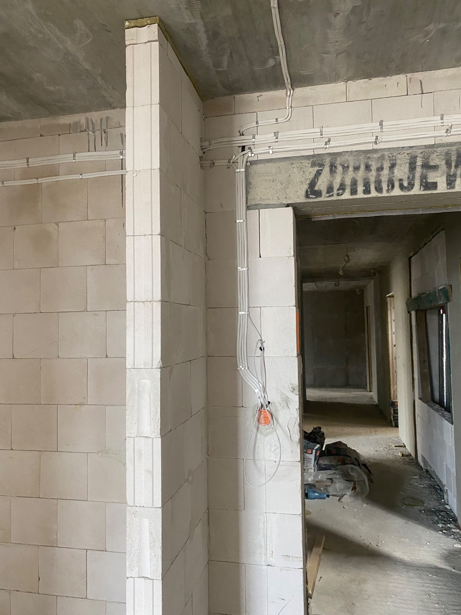 Wewnętrzne ściany z białych bloczków w surowym stanie, z widocznymi przewodami elektrycznymi mocowanymi do sufitu i ściany, oraz wystającymi kablami ze ściany, widok na korytarz w głębi.