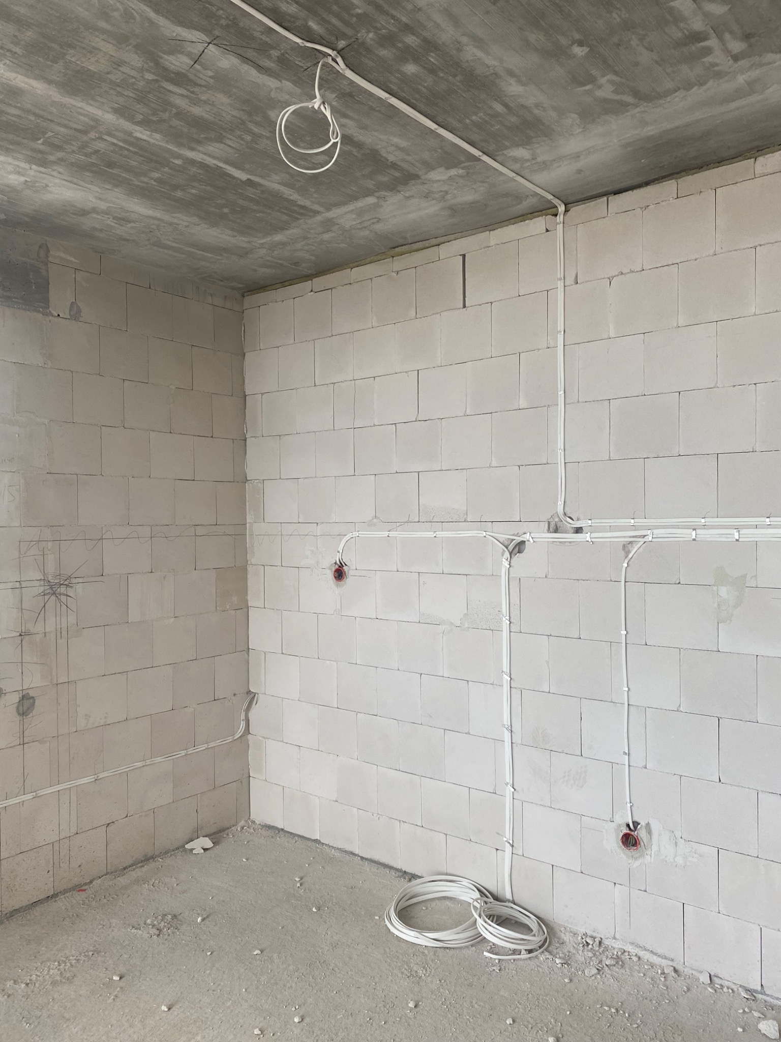 Surowy stan wnętrza z widoczną instalacją elektryczną: przewody prowadzone po ścianach z betonu komórkowego, gniazda w puszkach, pętle kabli zwisające z sufitu i leżące na podłodze.