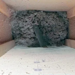 Wnętrze poddasza z widoczną warstwą izolacji z wełny mineralnej, przez którą wystaje czarna rura wentylacyjna; widoczne drewniane elementy konstrukcyjne poddasza.