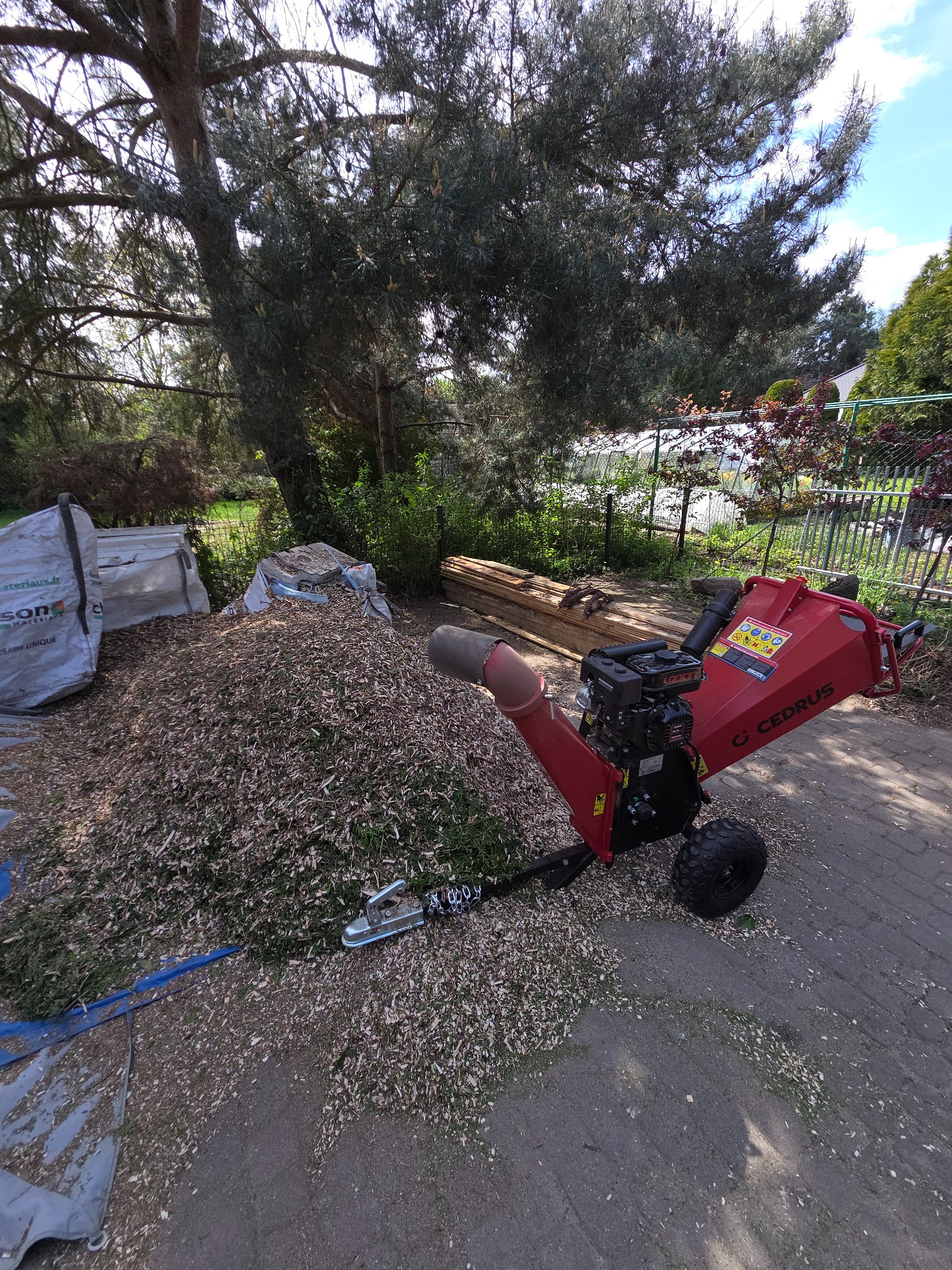 Czerwony rębak do gałęzi Cedrus obok sterty zrębków w ogrodzie, worki z materiałami w tle. Widoczne drzewa i fragment szklarni.