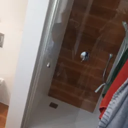 Szklana kabina prysznicowa z drewnopodobną ścianą, widoczne chromowane elementy armatury i wiszące ręczniki w odcieniach szarości, czerwieni i zieleni.