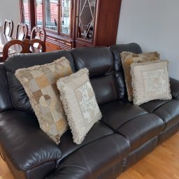 Agnieszka Kwiatkowska - Ciemnobrązowa skórzana sofa z dekoracyjnymi poduszkami w odcieniach beżu i brązu, widoczny fragment jadalni z drewnianymi meblami w tle.