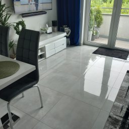 Agnieszka Kwiatkowska - Wypolerowana, jasnoszara podłoga z płytek ceramicznych, odbijająca światło, z fragmentem nowoczesnego salonu z białymi meblami, szarymi krzesłami i balkonem z roślinami w tle.