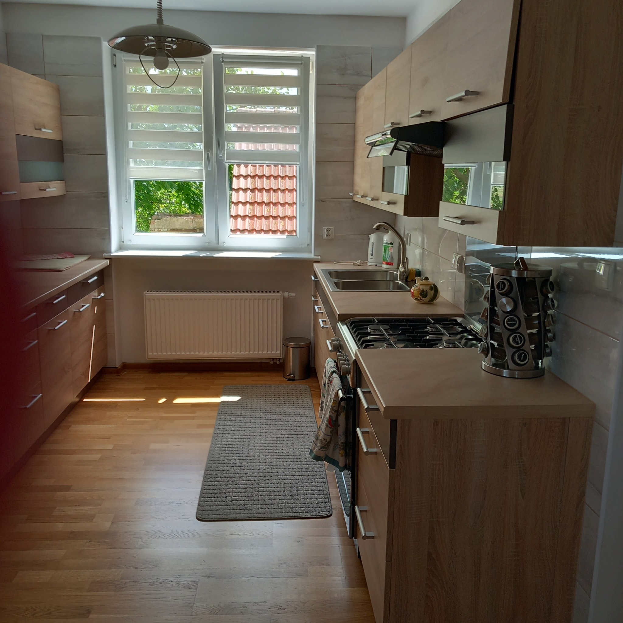 Jasna kuchnia z drewnianymi szafkami, widok na okno z roletami dzień-noc, grzejnik i dywanik podłogowy.