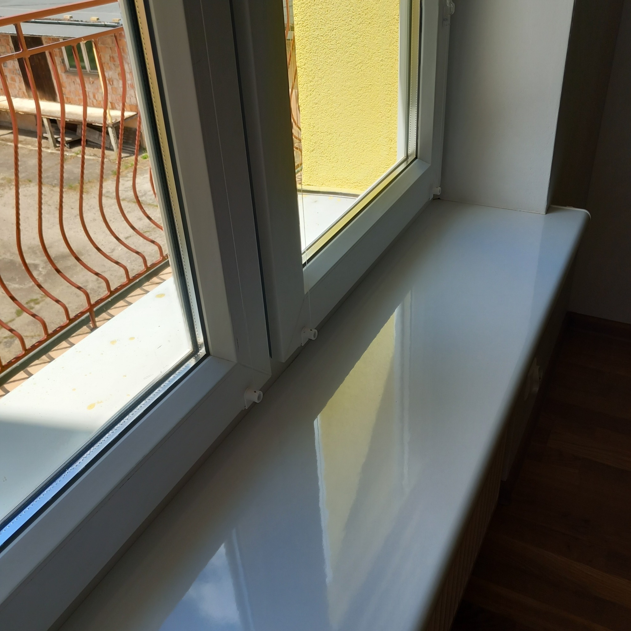 Błyszczący parapet z PCV po umyciu, widok z góry na okno z widoczną, zewnętrzną, żółtą ścianą budynku i balkonem z metalową balustradą.