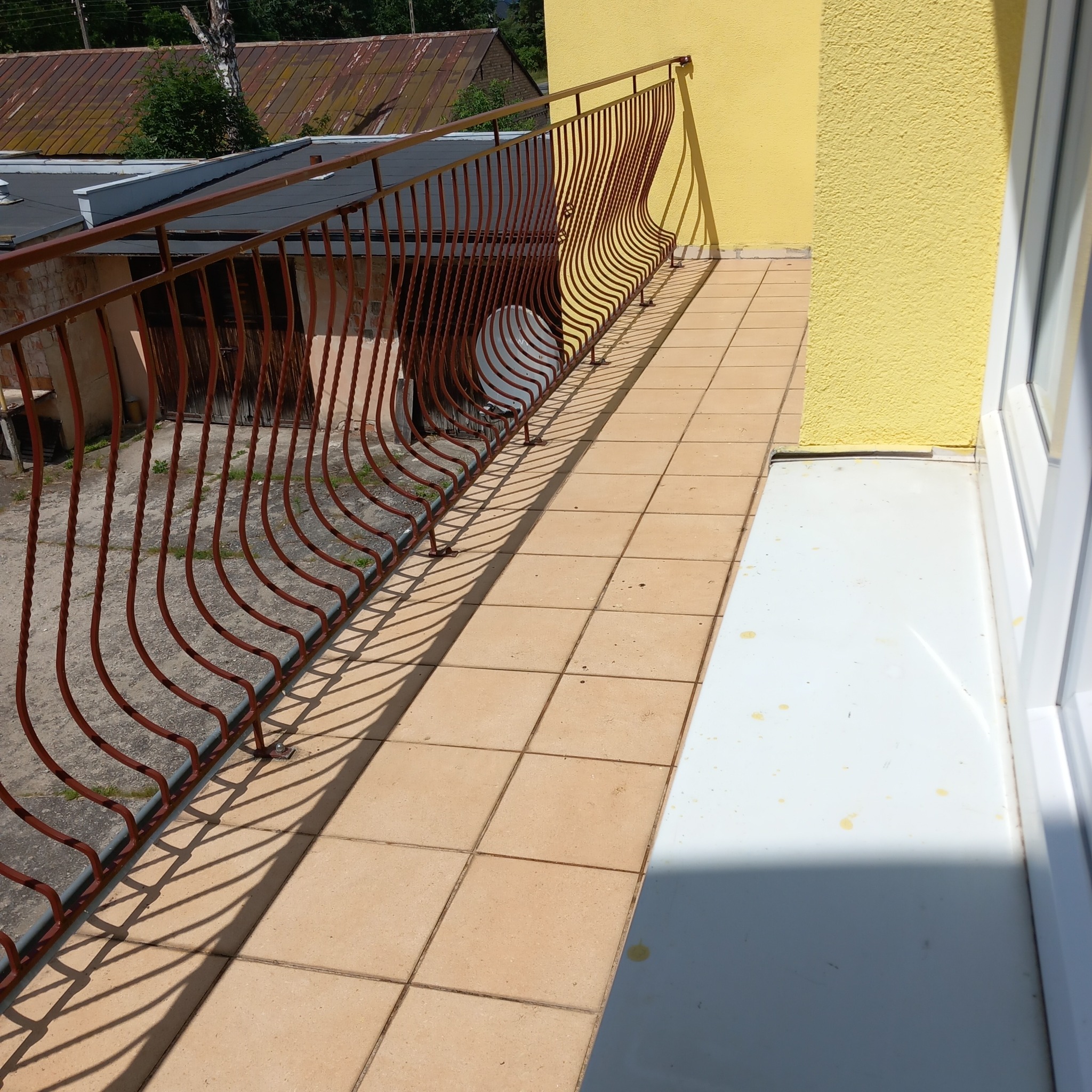 Balkon z brązową, dekoracyjną balustradą, wyłożony beżowymi płytkami, z żółtą ścianą i białym oknem, na którym widać drobne zabrudzenia.