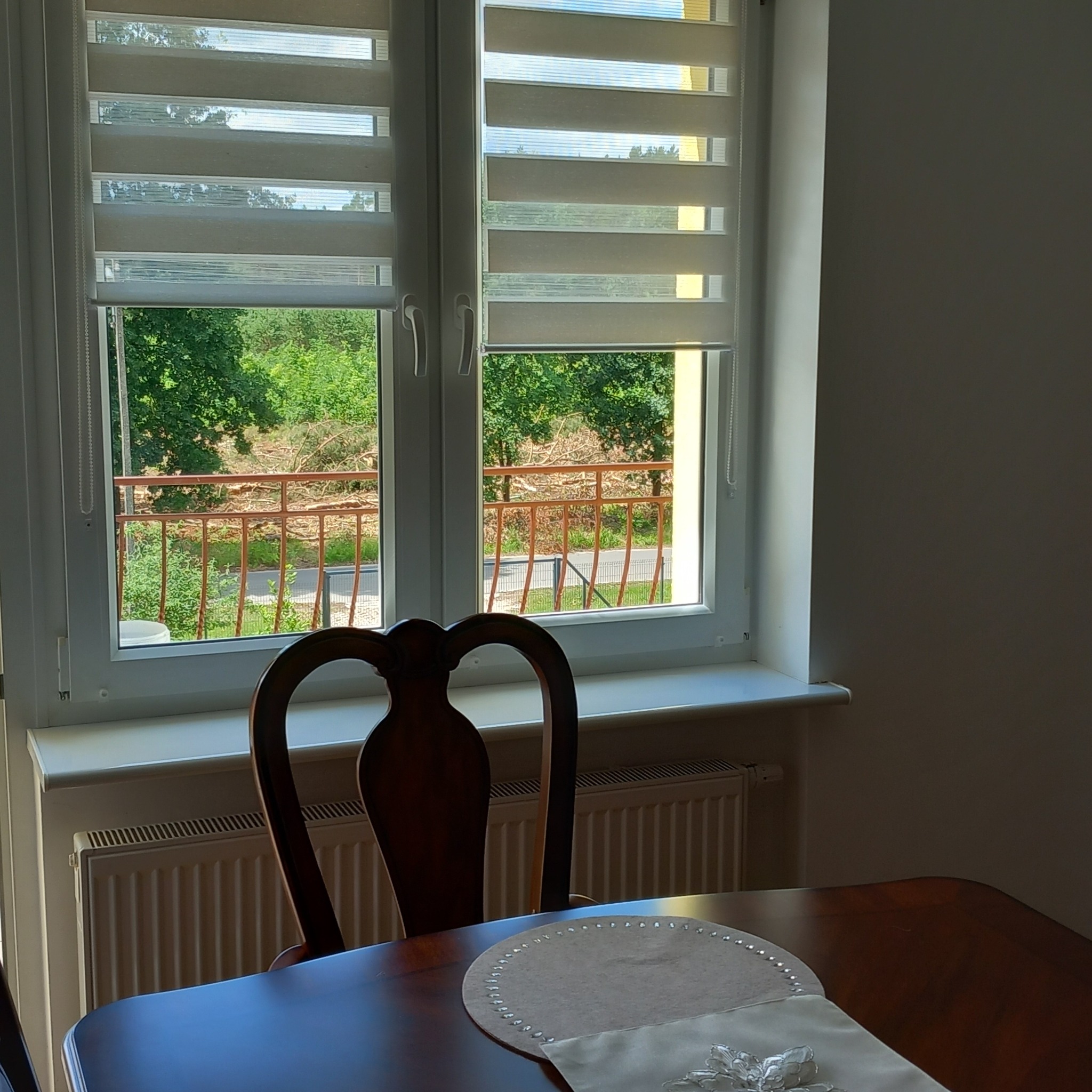 Wnętrze pokoju z drewnianym stołem, krzesłem z wysokim oparciem i oknem z żaluzjami, przez które widać zieleń drzew i balkon.