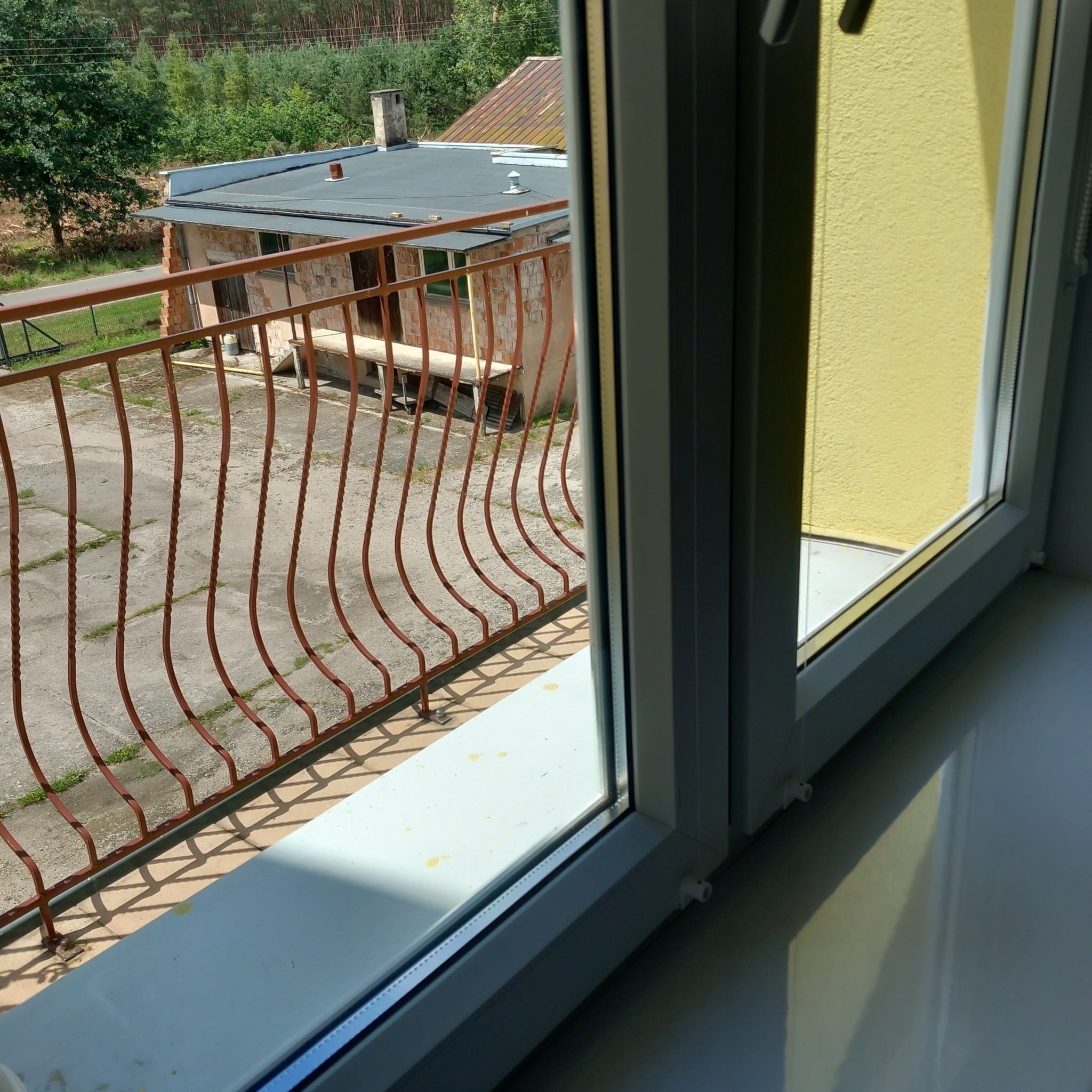 Widok z okna na balkon z brązową, metalową balustradą i budynek gospodarczy z cegły, w słoneczny dzień. Na parapecie widoczne drobne zabrudzenia.